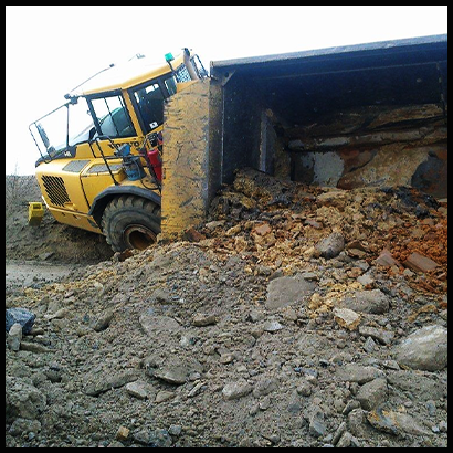 Articulated dump truck listing, with large mound of gravel spilled onto ground from tipped-over trailer.