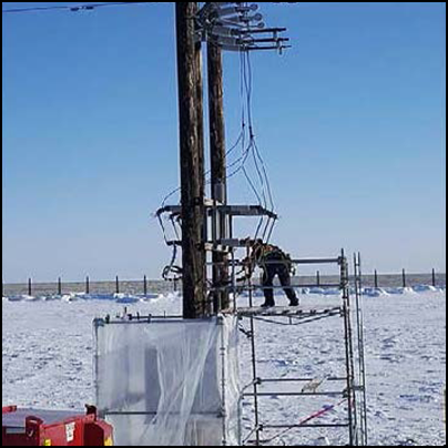 一名工人正站在约2米高的脚手架上，在电线杆组件和架空电线旁边搭建脚手架。 