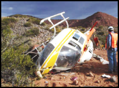 The crashed helicopter overturned with damage to the body and propeller. A worker wearing a protective hat and high visibility vest examining the helicopter. 