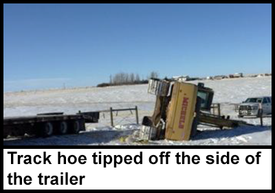 The tipped over track hoe lying next to the end of a trailer.
